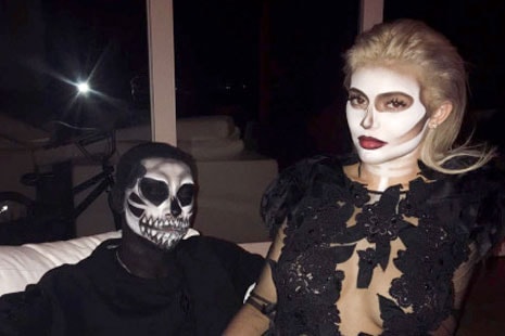 Kylie Jenner con vestido negro y cara pintada de blanco sentada en las rodillas de alguien vestido de negro y máscara de calavera