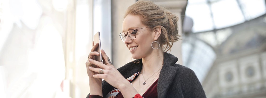 mejores apps de turismo para viajar Mujer con gafas y bolsas en el brazo mira hacia su teléfono móvil que sujeta en la mano