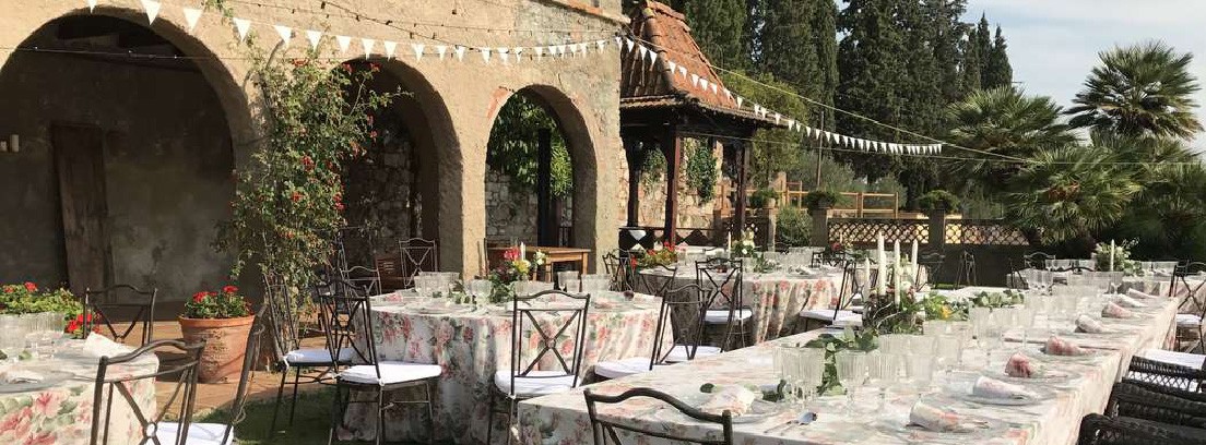Mesa alargada rodeada de mesas redondas con manteles de flores y sillas en jardín