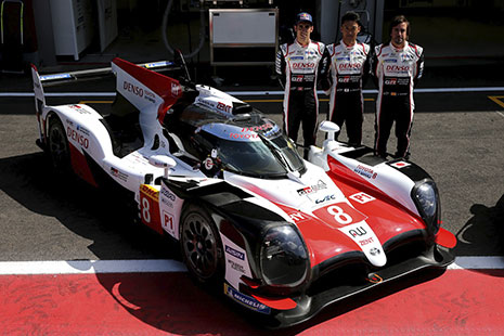 Coche de carreras blanco y rojo junto a él tres hombres con mono de piloto