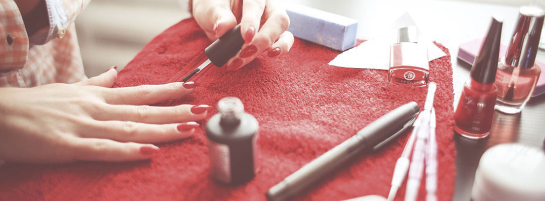 Mano sobre toalla roja con uñas rojas y sobre ella otra mano sujeta un pincel de pintauñas