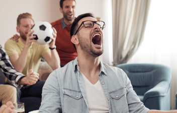 Un grupo de amigos en un salón celebre un gol de su equipo de futbol. Uno de ellos porta un balón de fútbol