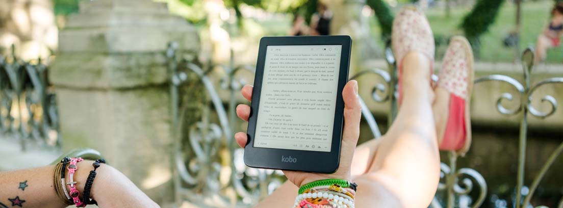 Bookchoice: 8 libros al mes Manos con pulseras sujetan un ebook sobre piernas cruzadas, extendidas sobre una barandilla