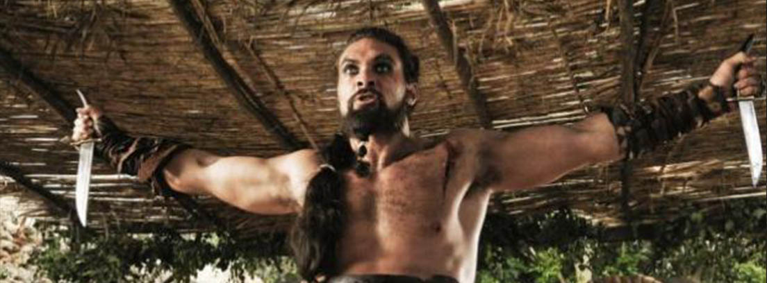 Momoa como Khal Drogo con coleta larga, torso desnudo y brazos en cruz con cuchillos.