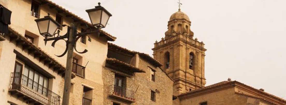 Pueblos más bonitos de españa Mirambel en Teruel Resto de muralla de piedra junto a fachada de casa y delante de torre de la iglesia de Mirambel (Teruel).