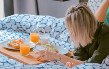 Mujer tumbada sobre una cama de hotel con una bandeja con comida.