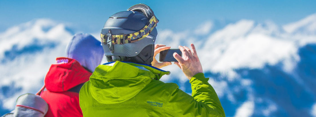 Dos personas en la nieve enfocando su móvil hacia montaña nevada.