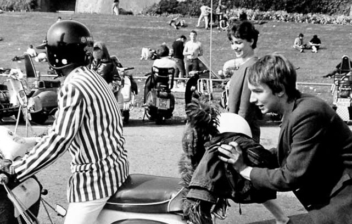 Varios mod empujan una scooter sobre la que hay otro hombre montado.