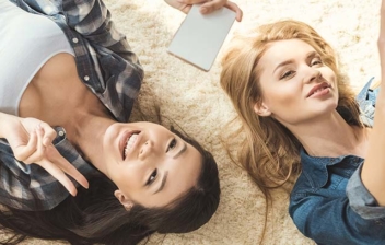 Dos mujeres tumbadas sobre alfombra de pelo mirando hacia teléfonos móviles